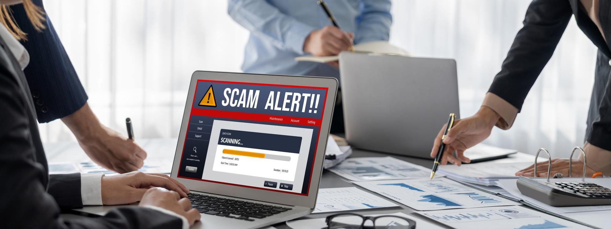 Laptop screen shows SCAM ALERT!! during business meeting.