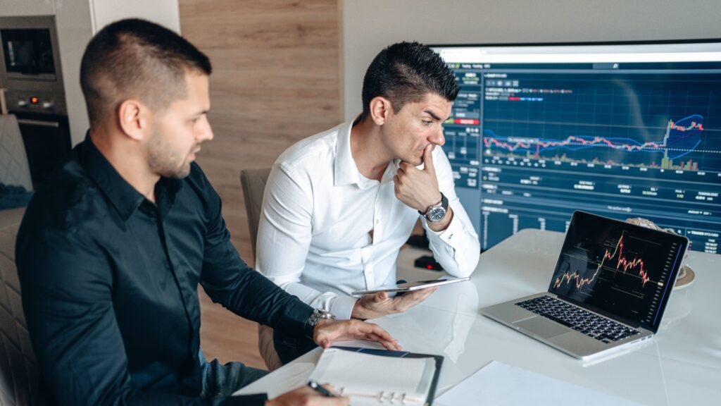 Two businessmen sitting at a desk analyzing stock charts on laptops and tablets, representing collaborative investment decisions